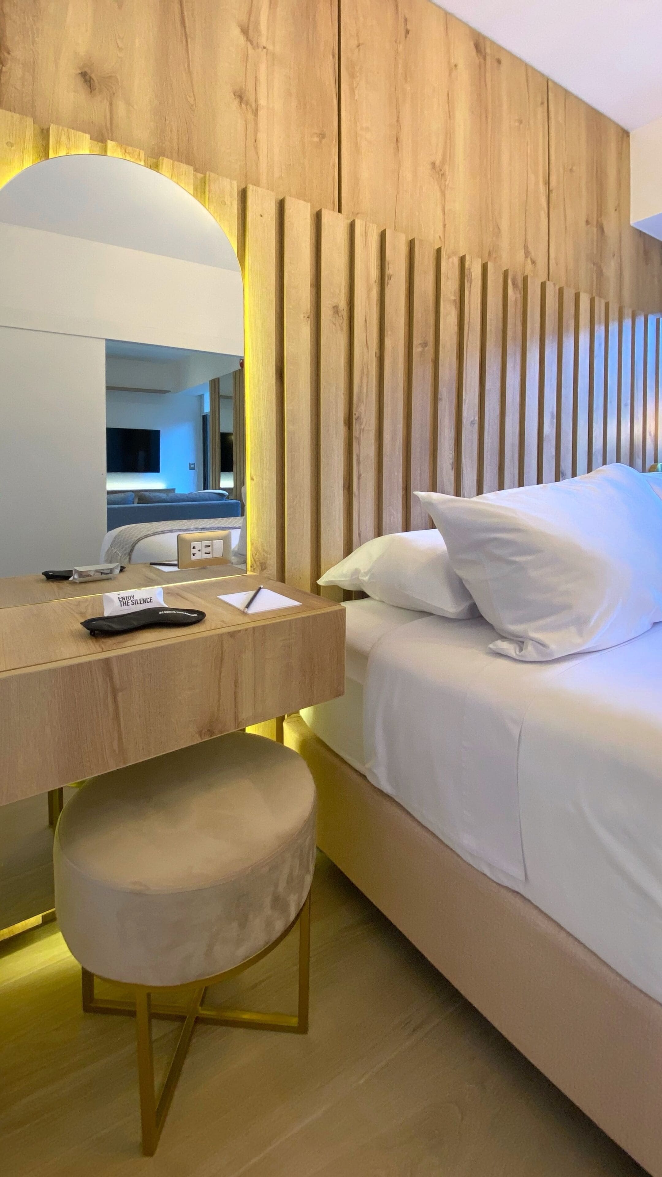 Een moderne hotelkamer met een houten hoofdeinde, een nachtkastje met een lamp, een rond krukje en een bed met witte lakens.