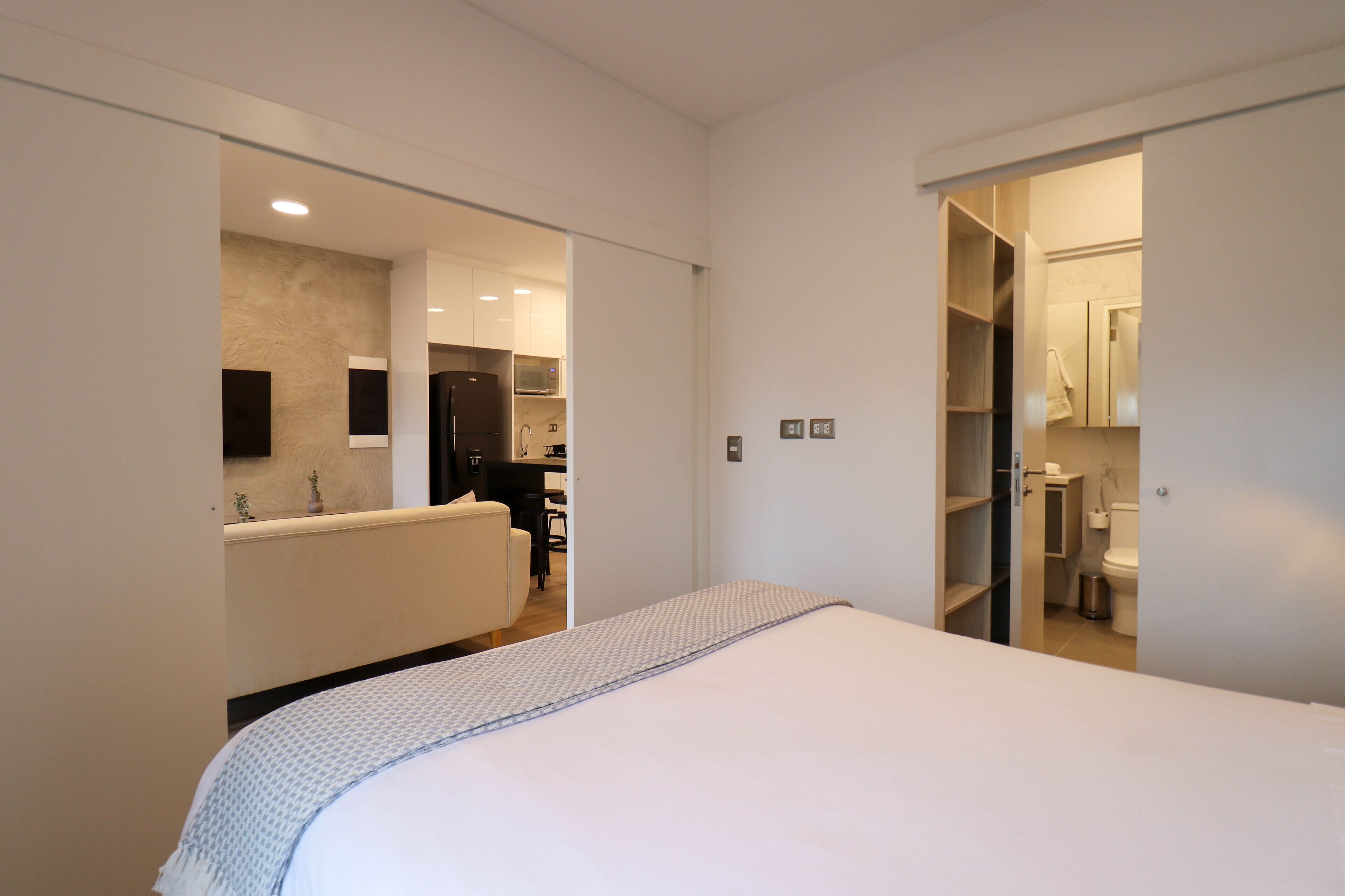 Una habitación de hotel moderna con una cama grande, un baño con inodoro y lavamanos, y una zona de kitchenette.