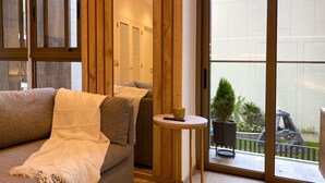 A modern living room with a sofa, a small round table with a bowl and a remote control, and a view of a balcony with plants and outdoor furniture.