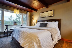A neatly made bed with a quilted white comforter, a wooden headboard, a bedside table with a lamp, and a window with a view of trees. - Columbine Cabin at Highland Meadows - Black Hills Retreat (Hot Springs)