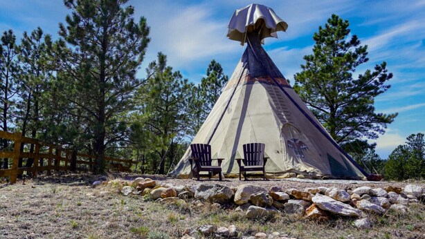 En tipi med to Adirondack stole foran, omgivet af sten og grantræer.