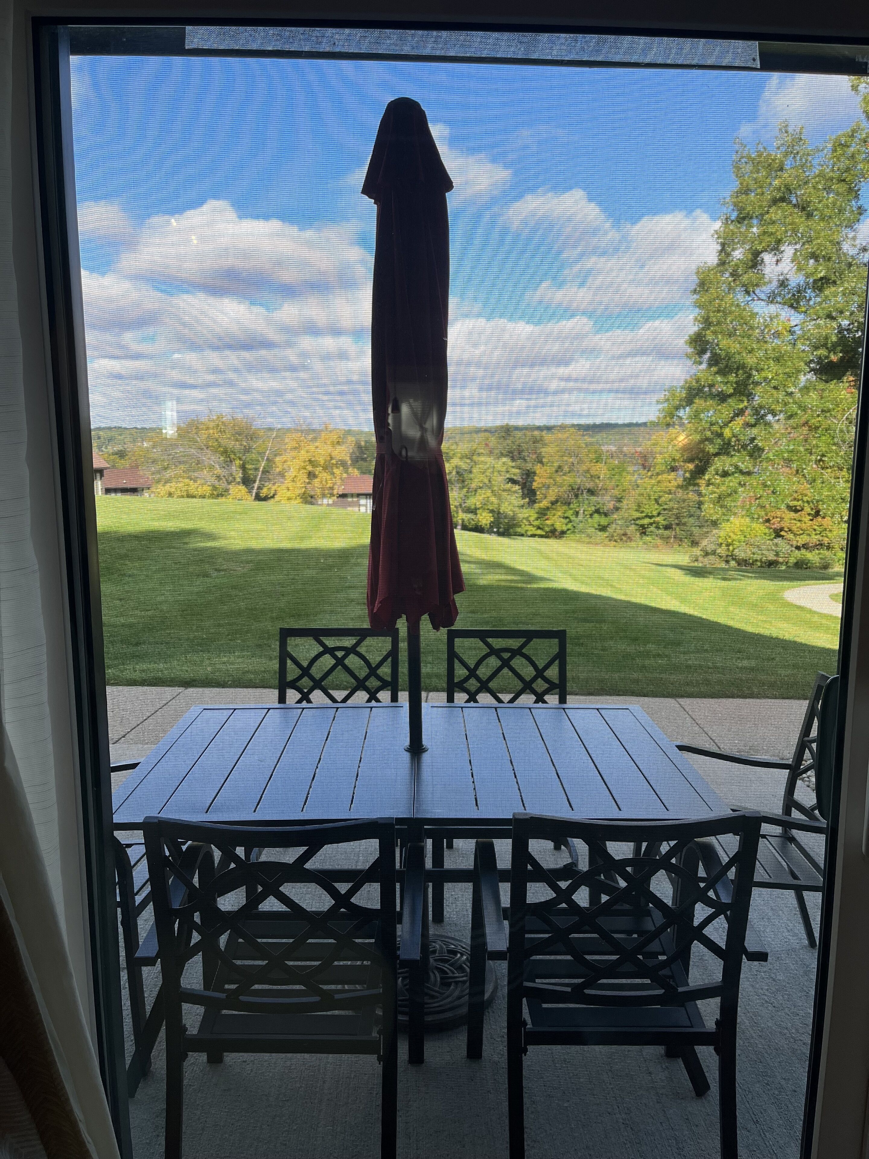 Une terrasse avec une table et des chaises, un parasol, et une vue sur un espace verdoyant avec des arbres et un bâtiment au loin.