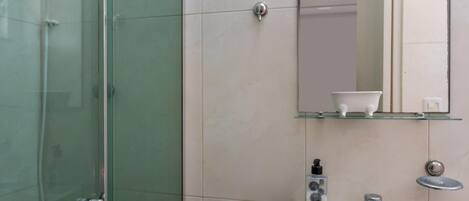Un baño con cabina de ducha de vidrio, lavamanos pedestal, espejo y un estante con un objeto blanco.