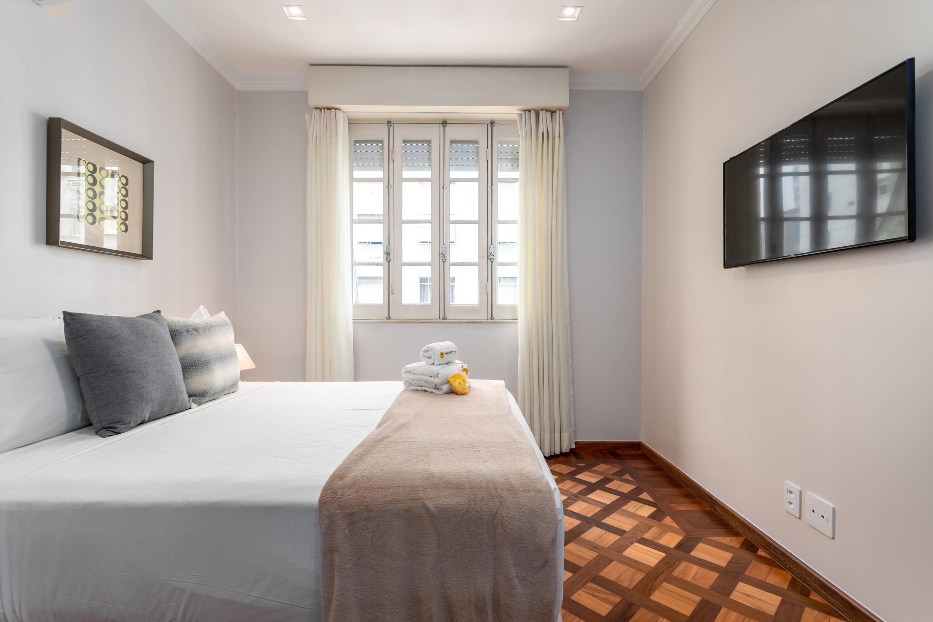 A hotel room with a bed, a TV mounted on the wall, a window with curtains, and a patterned floor.