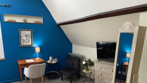 A room with a blue accent wall, a wooden desk with a chair, a black armchair, a dresser with drawers, a small round table with a lamp, a television, and a radiator. - La Maison de Triboulet, Chambres et Table D'hôtes, Extingo, Extingo (Pocé-sur-Cisse)