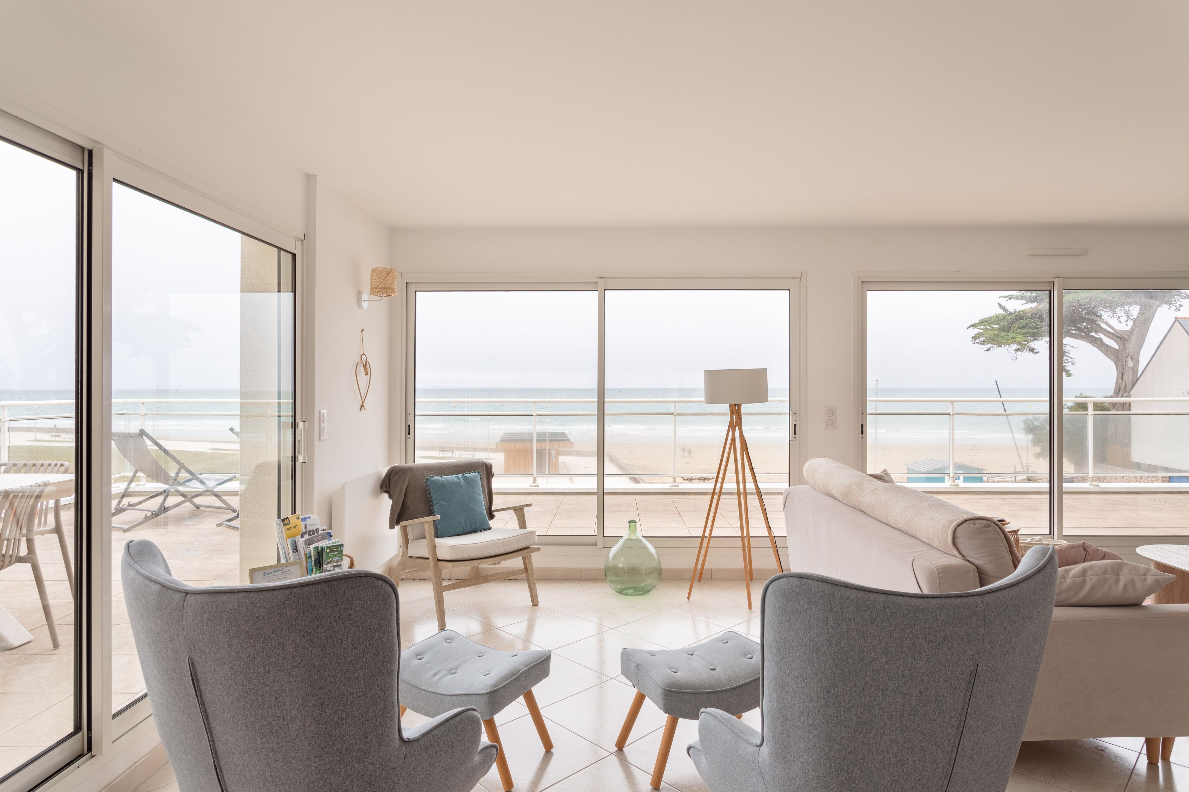 A modern living room with a view of the beach, featuring a sofa, two armchairs, a floor lamp, and a small table with magazines.