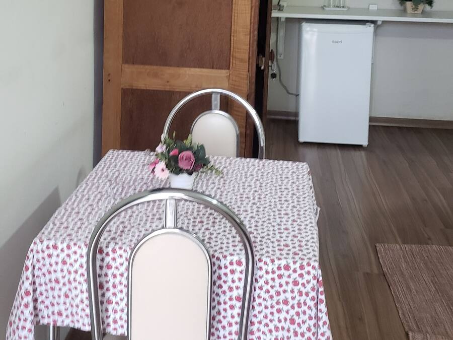 Uma sala de jantar com uma mesa posta para dois, uma porta de madeira, uma geladeira branca e um pequeno vaso com planta.