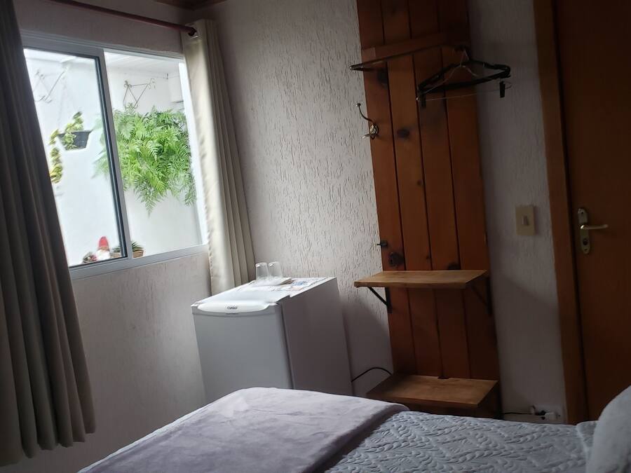 A bedroom with a bed, a small table, a washing machine, a wooden wardrobe, a window with curtains, and a view of greenery outside.
