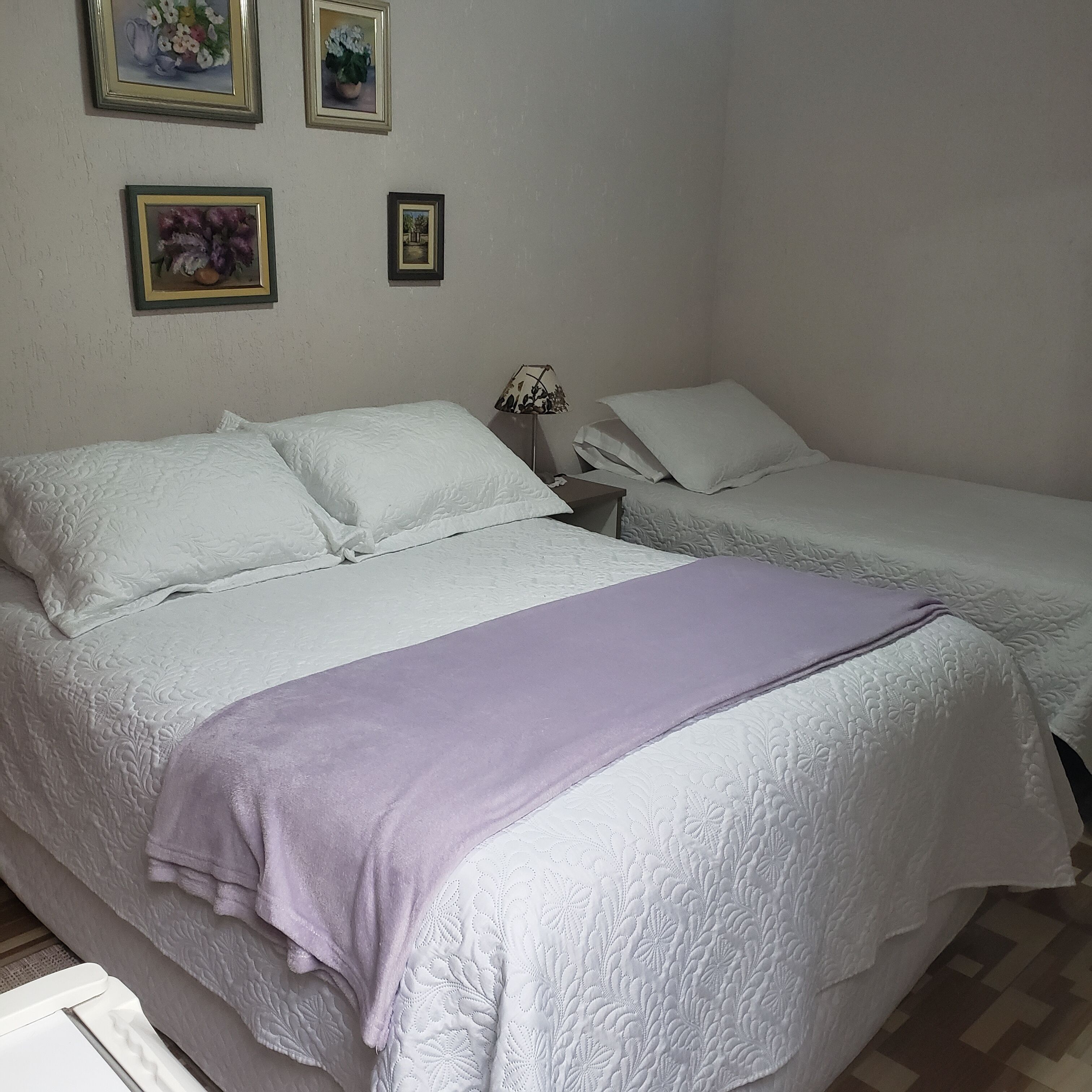 A bedroom with two beds, a nightstand, and a lamp. There are three framed pictures on the wall.