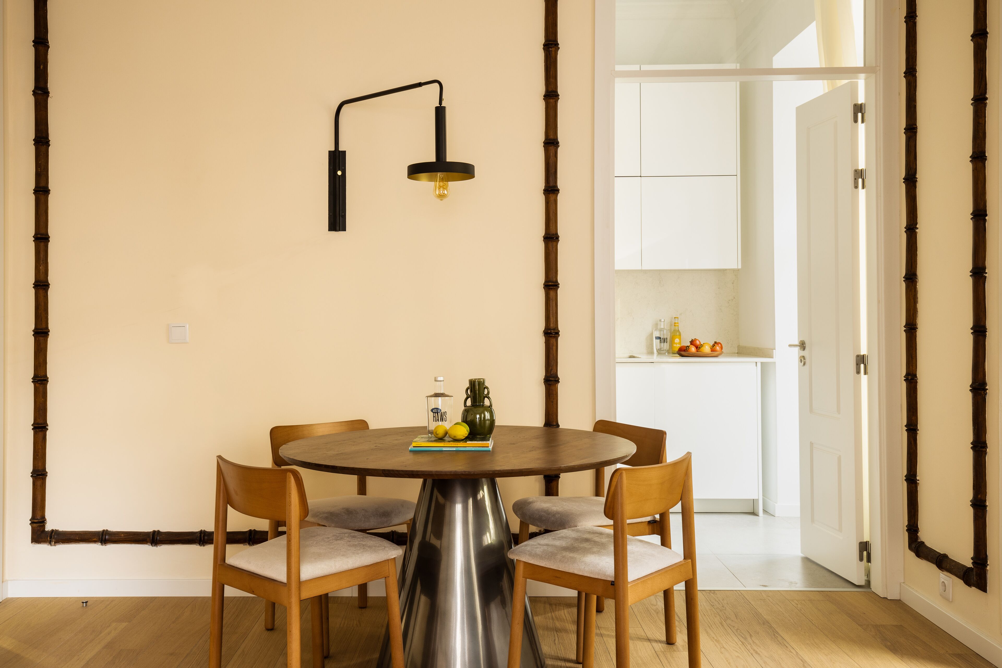 Uma sala de jantar com uma mesa redonda, quatro cadeiras, luminária fixada na parede e cozinha visível através de uma porta aberta.