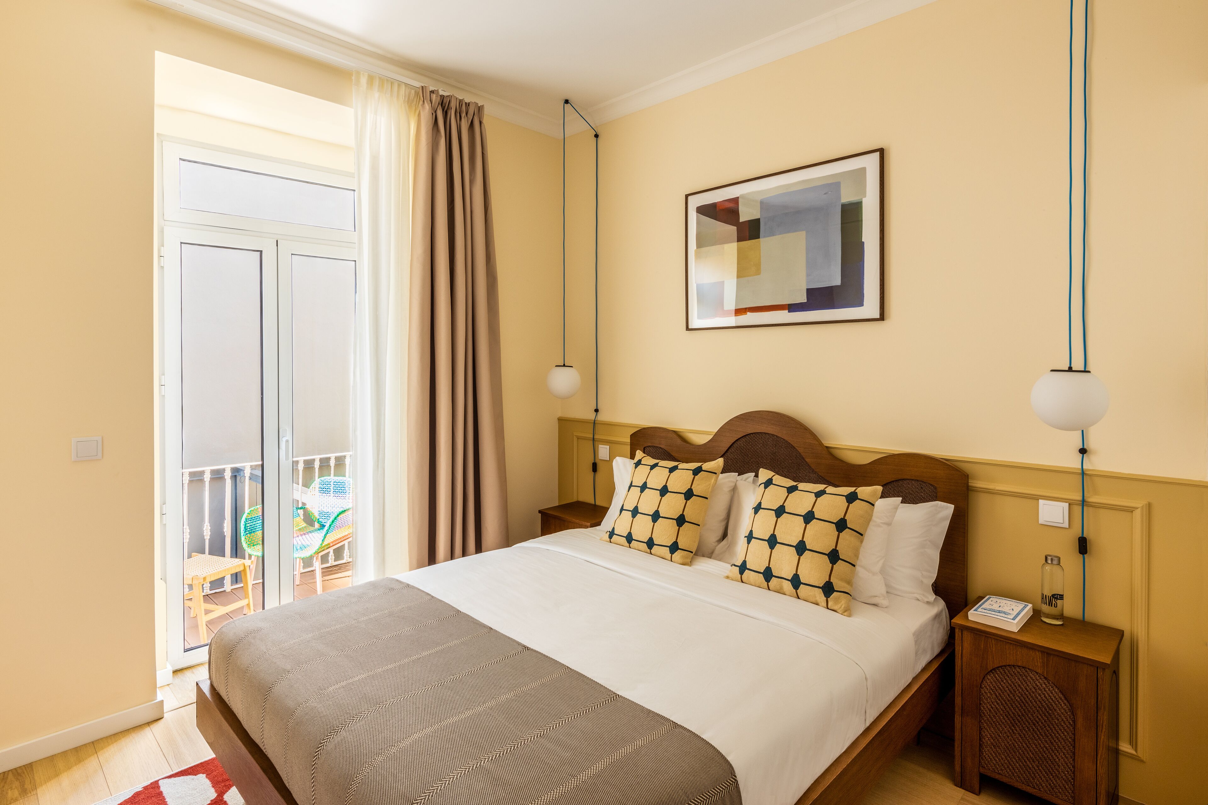 A hotel room with a large bed, a wooden headboard, a framed abstract painting, a nightstand, and a view of the balcony.