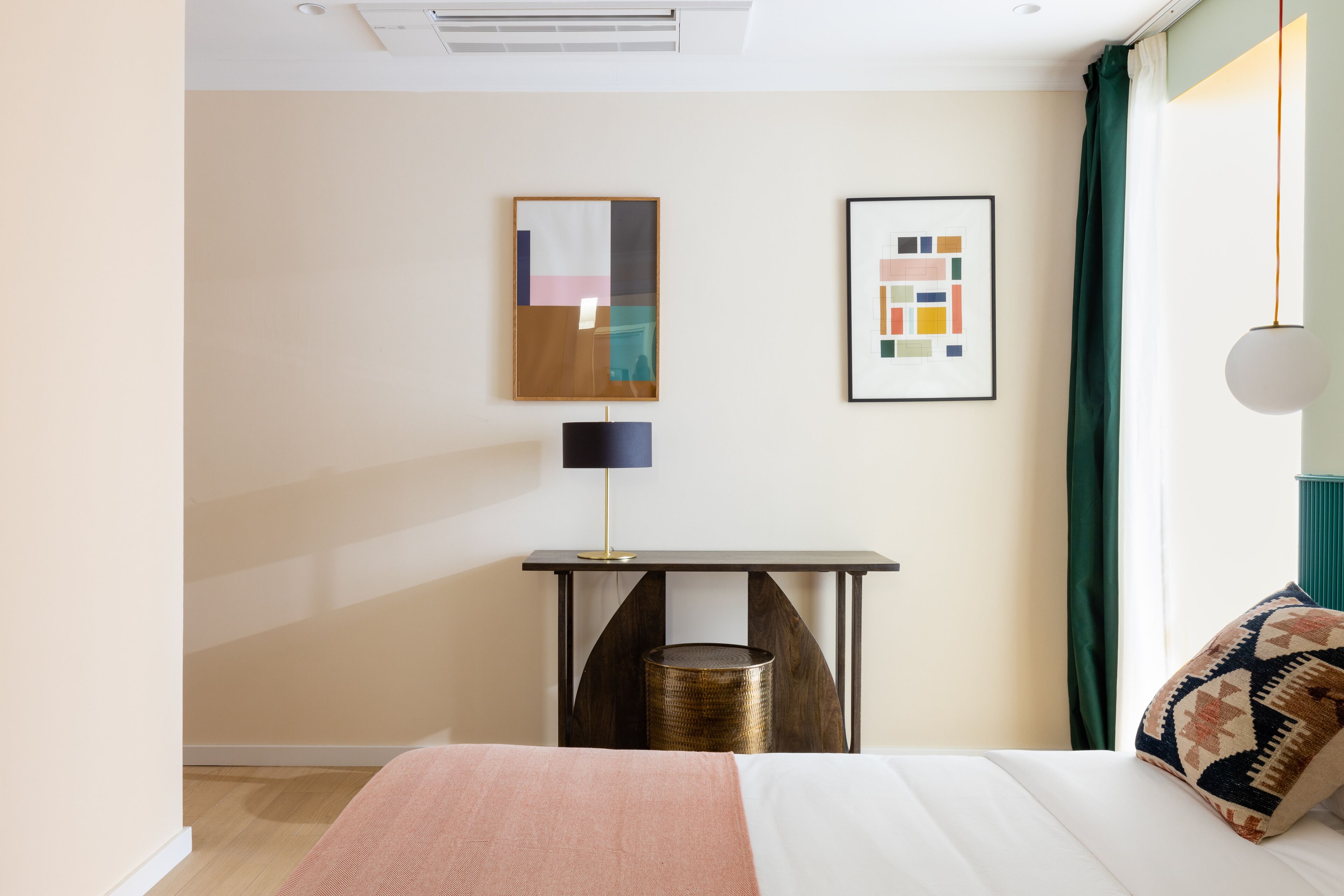 Habitació d'hotel amb un llit, una taula de nit amb una làmpada, un safataire i dues obres d'art amb marc a la paret.