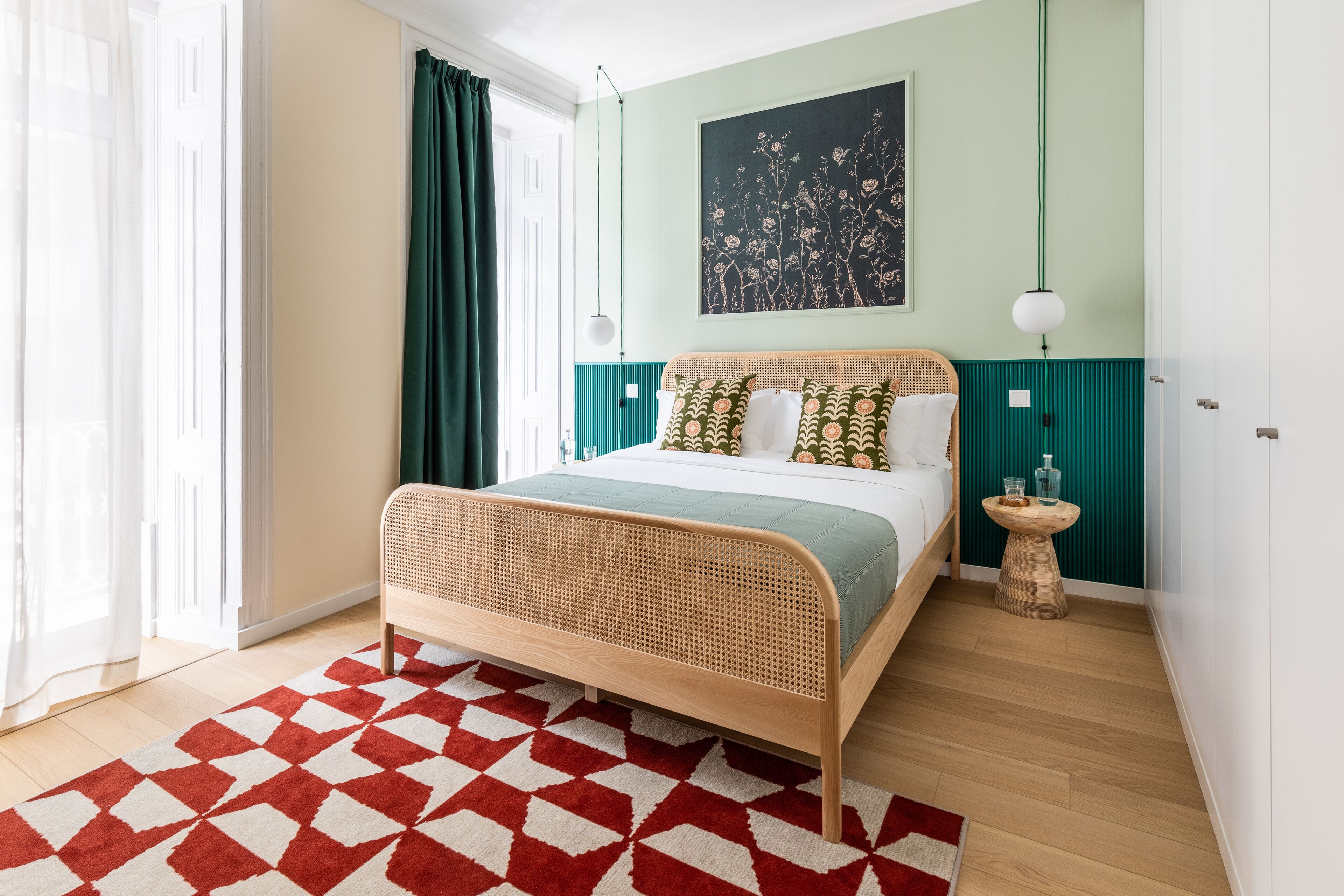 Un dormitorio con cama de ratán, alfombra estampada con motivos geométricos, pared de acento color turquesa, lámpara de pared y una pequeña mesita de noche de madera.