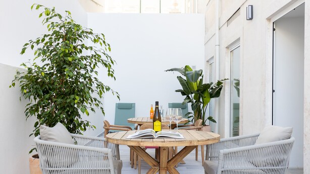 Una mesa de madera redonda con sillas de mimbre, rodeada de plantas en macetas, en un espacio interior luminoso.