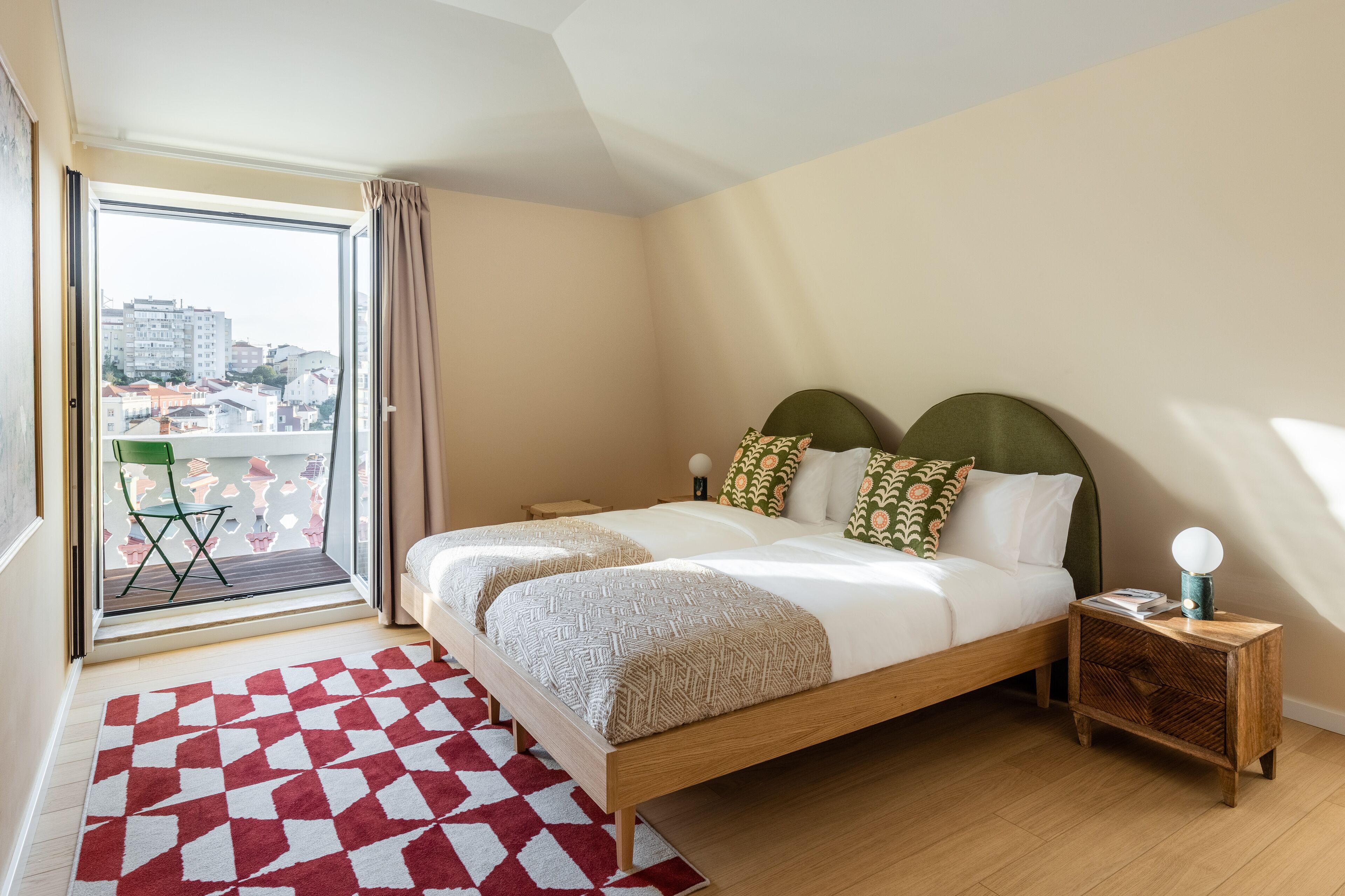 Un dormitorio con una cama grande, una mesita de noche, una alfombra roja y blanca, y vistas a edificios a través de una ventana.