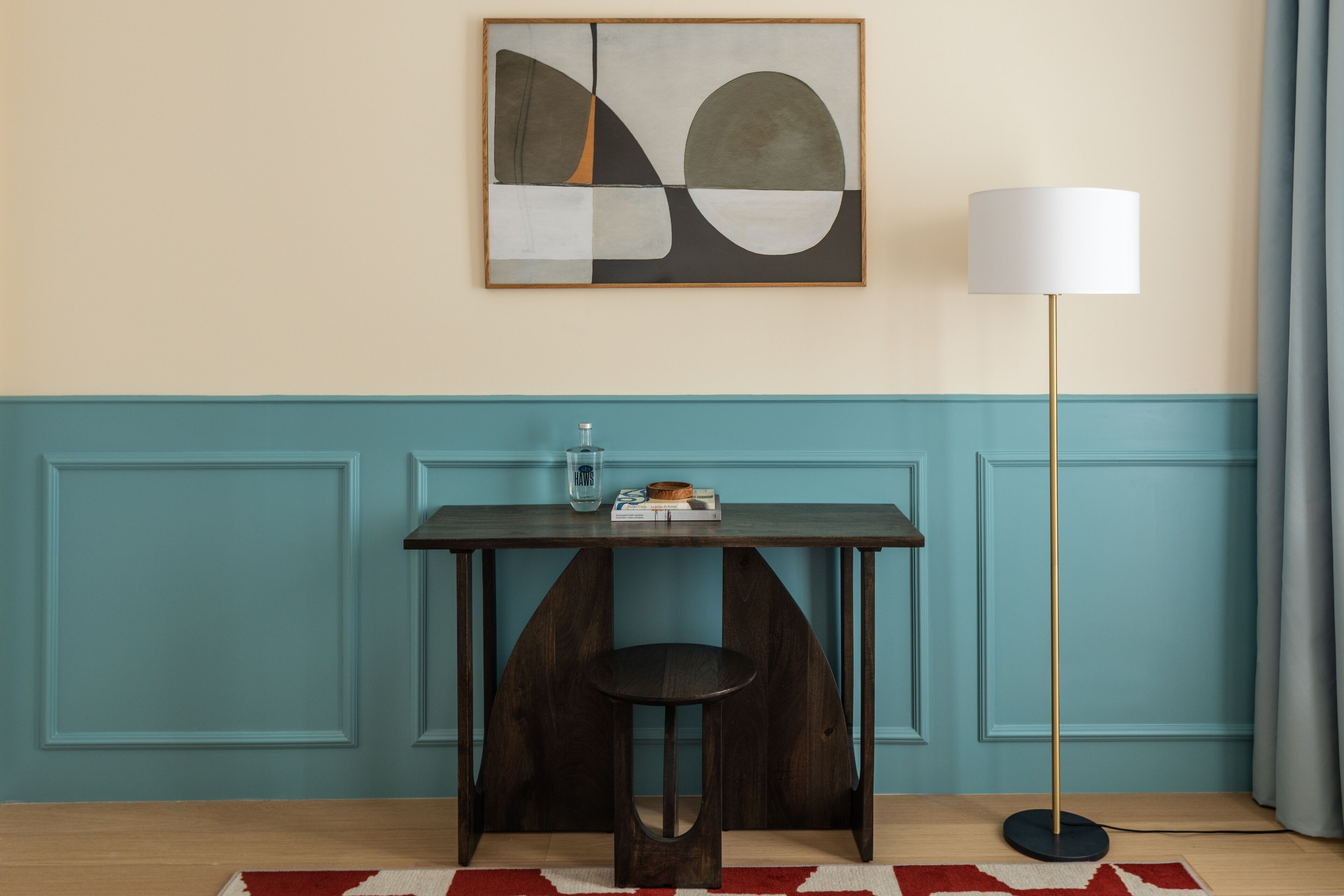 A room with a dark wooden table, a single chair, a floor lamp, and a wall-mounted abstract painting.