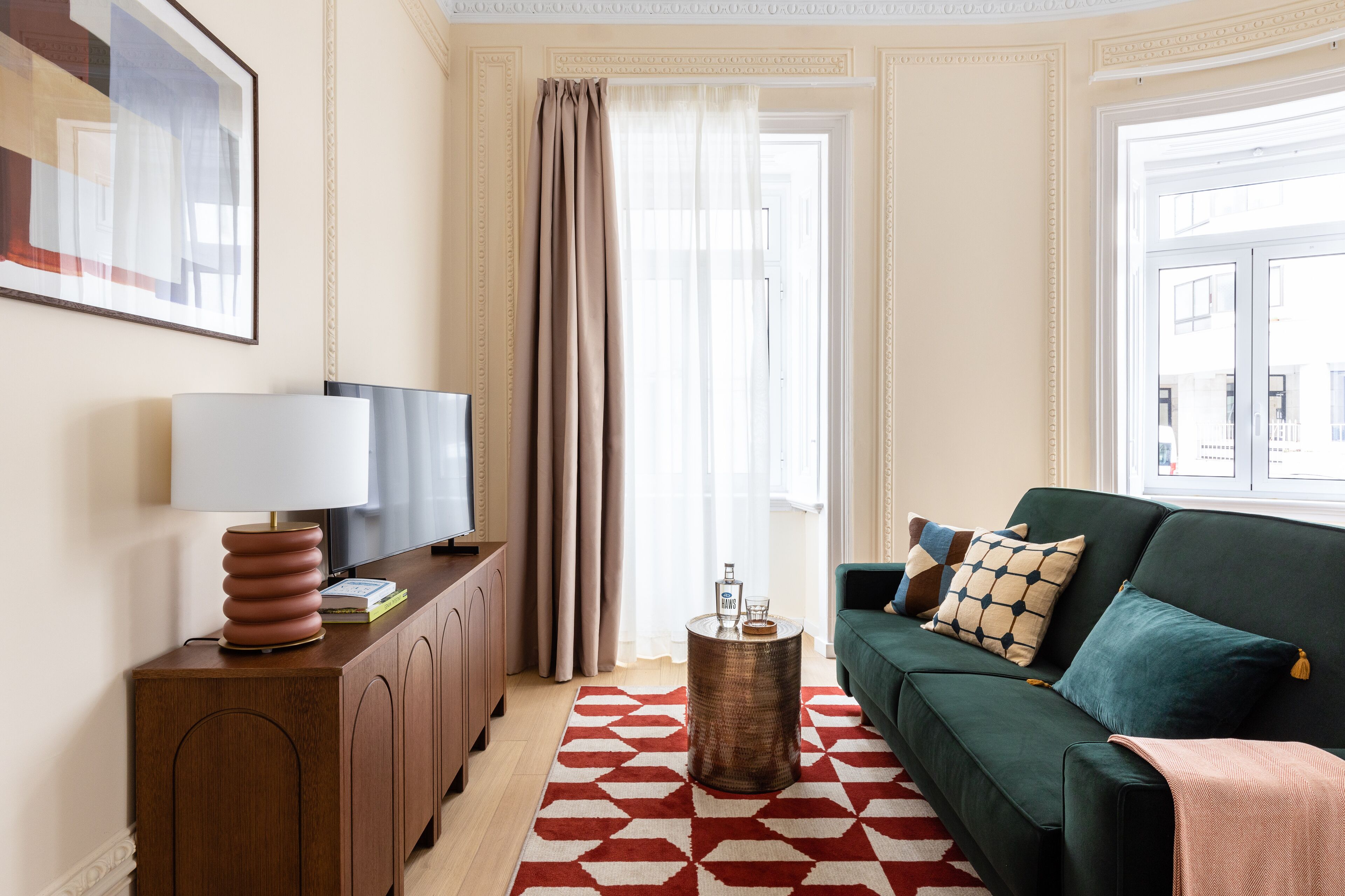 A living room with a green sofa, a wooden cabinet, a red and white patterned rug, and a large window with curtains.