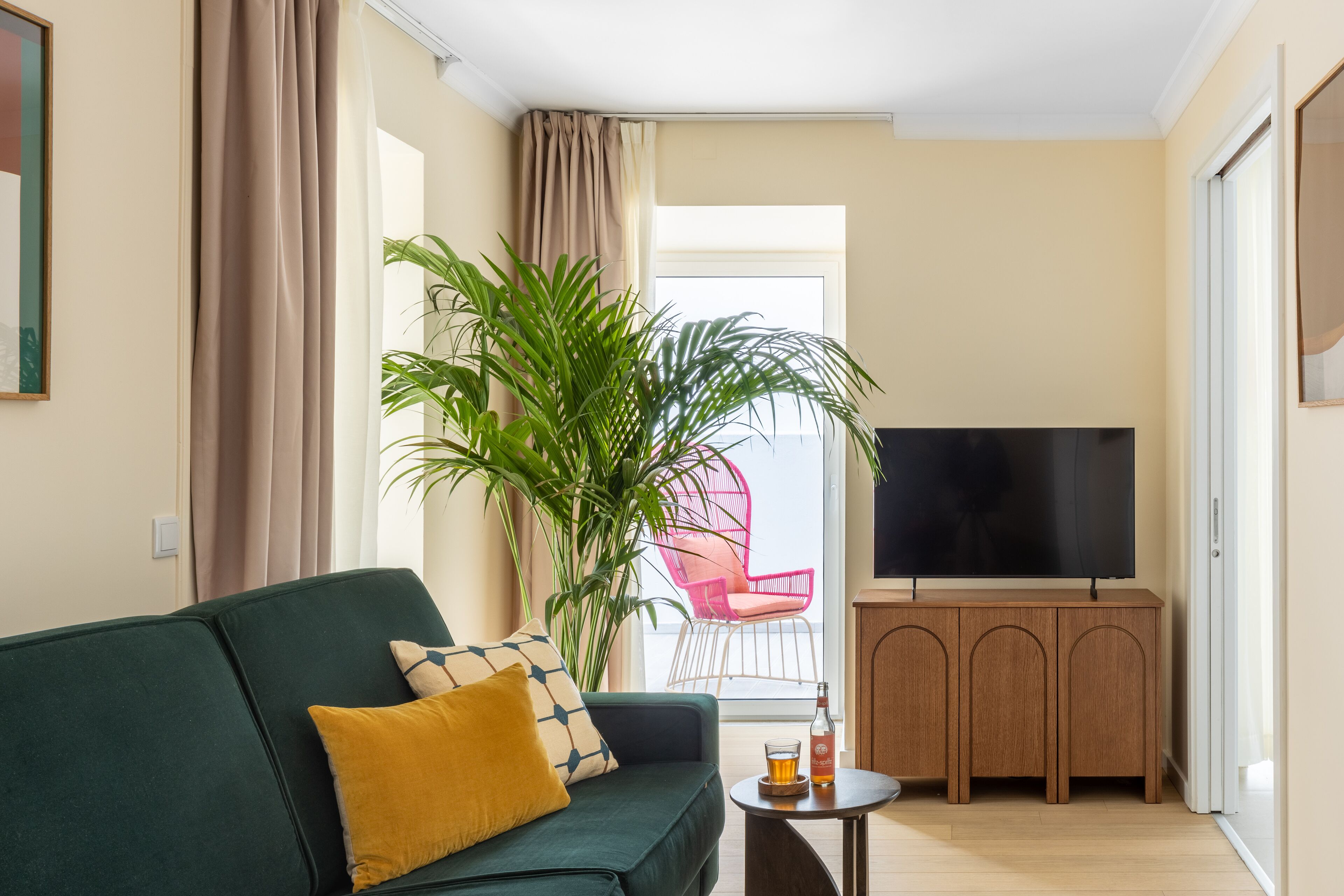 Sala de estar com sofá verde, uma mesa pequena com uma garrafa, televisão em um móvel de madeira e uma planta grande.