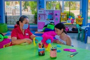 Two individuals seated at a table with colorful toys and art supplies, in a room with a play area in the background.