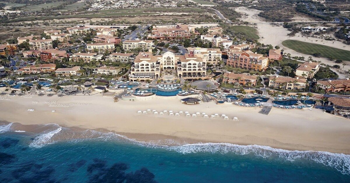 Vista aérea de un complejo turístico costero con varias edificaciones, una playa de arena y varias piscinas.