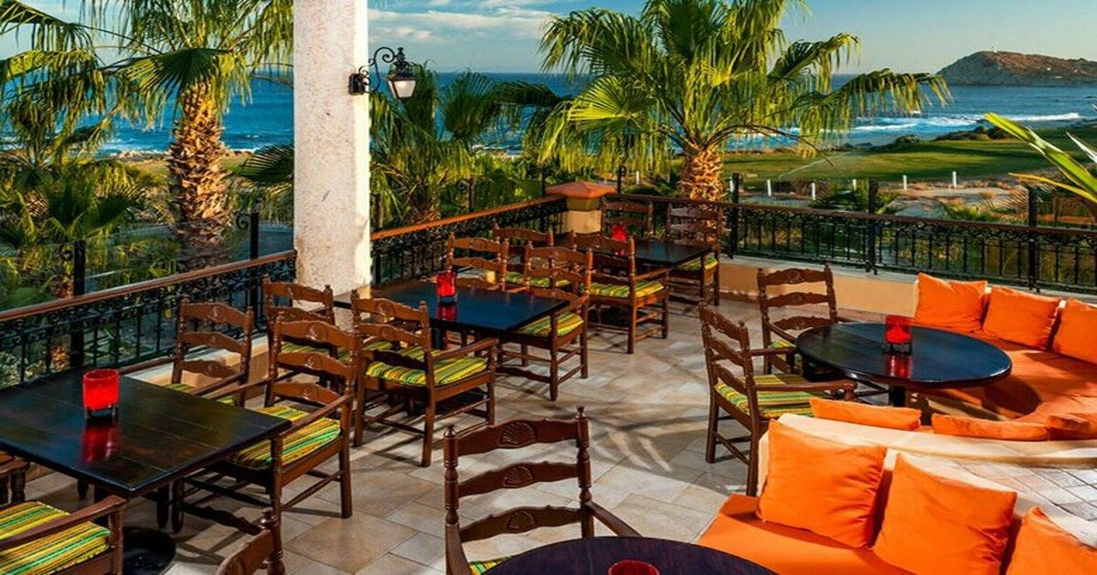 An outdoor seating area with tables and chairs, a view of the ocean, and palm trees.