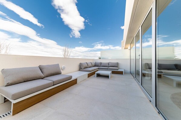 A modern outdoor seating area with a sectional sofa and a coffee table, large windows, and a clear sky.