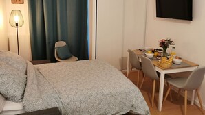 A hotel room with a bed, a desk with breakfast items, a TV, and a window with curtains. - Cosy Apartment  37m2 in the heart of Natural Park of Pyrenees (Aulus-les-Bains)