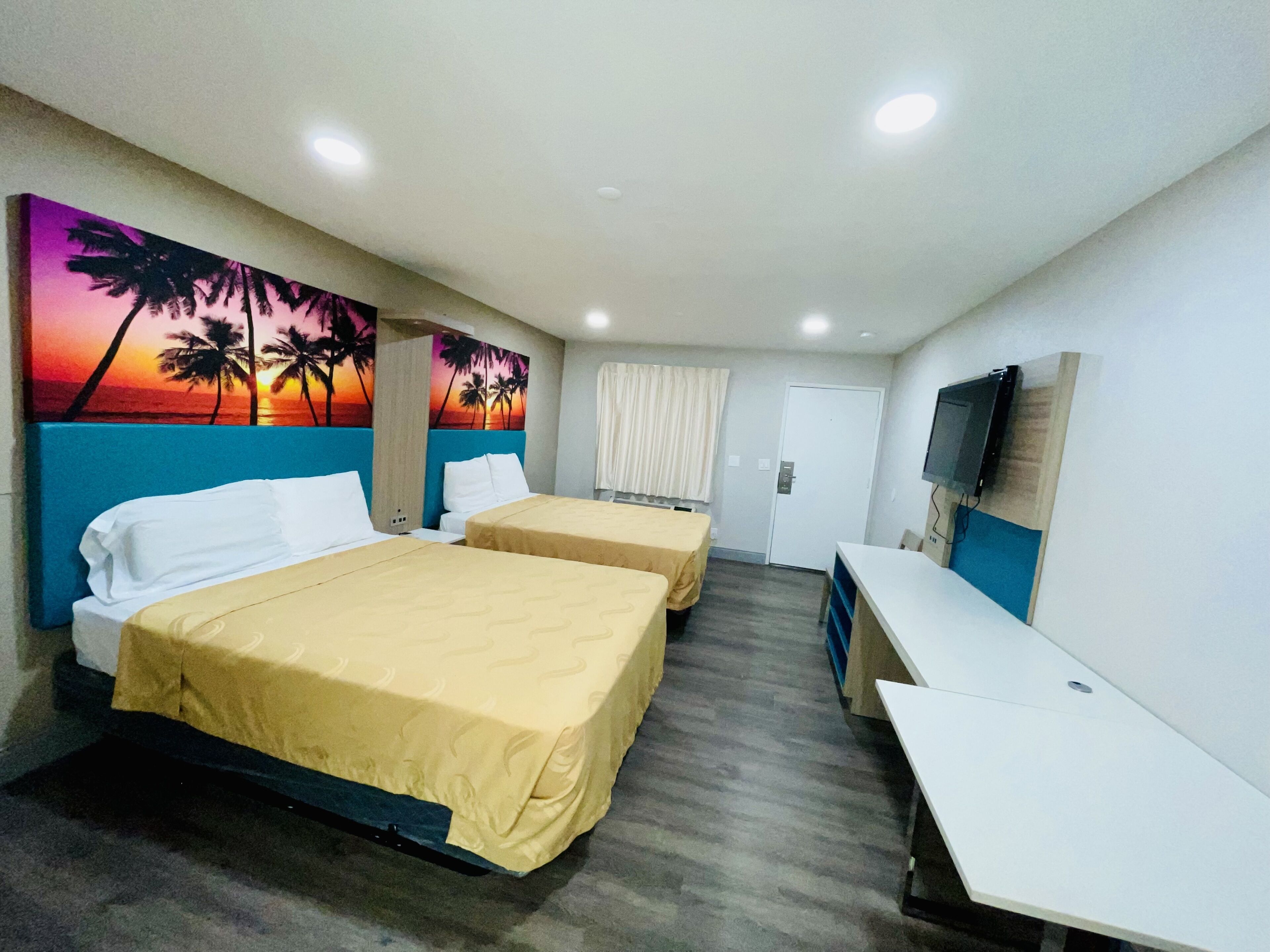 Habitación de hotel con dos camas, un escritorio, un televisor y un cuadro con un atardecer y palmeras.