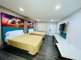 Habitación de hotel con dos camas, un escritorio, un televisor y un cuadro con un atardecer y palmeras.
