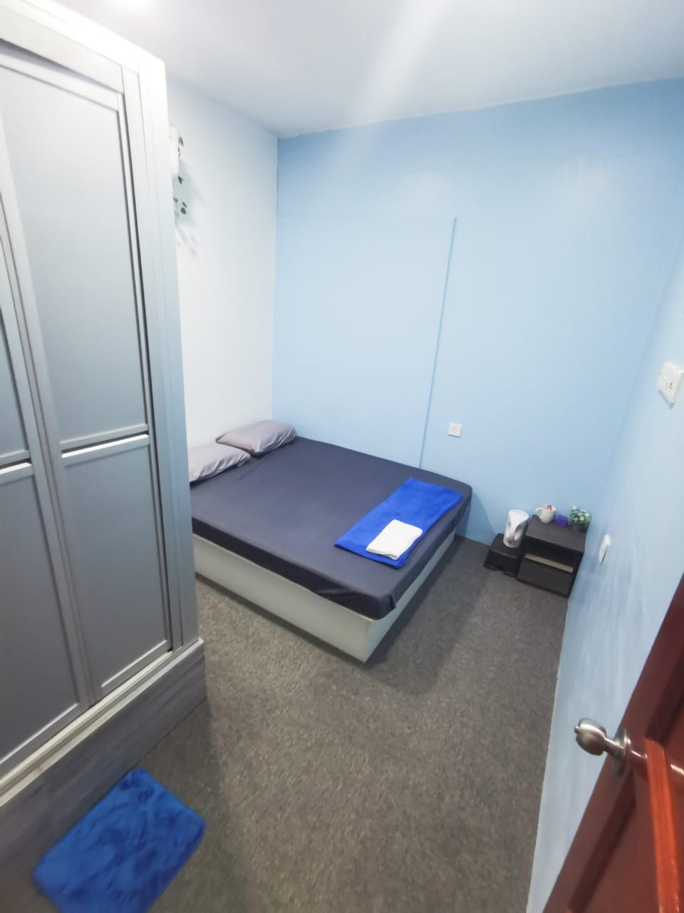 A small, single-bed room with a grey wardrobe, a blue mat, and a small table with a cup and a plant.