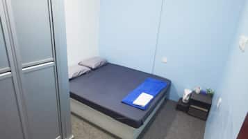 A small, single-bed room with a grey wardrobe, a blue mat, and a small table with a cup and a plant.