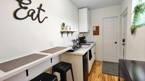 A compact kitchen with a white countertop, black stools, a coffee machine, and a wall-mounted shelf with a potted plant. - Cozy Studio with Kitchenette Attached (Davenport)