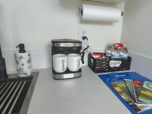 A kitchenette with a coffee maker, two mugs, a bottle of hand soap, a basket of bottled water, and a welcome mat. - Cozy Studio with Kitchenette Attached (Davenport)