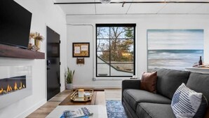 Un salon moderne avec une cheminée, une grande fenêtre donnant sur les arbres, un canapé gris et un téléviseur à écran plat fixé au mur.