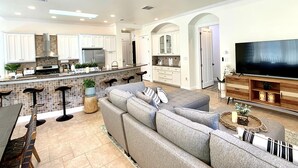A spacious living area with a large gray sectional sofa, a kitchen with white cabinets, a bar area, and a flat-screen TV on a wooden stand. - New Vista | Chef Kitchen, Golf Course Views (San Antonio)