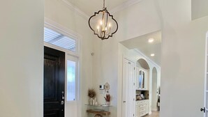A spacious hallway with a black door, a glass console table with decorative items, a white cabinet with glass doors, and a round ottoman. - New Vista | Chef Kitchen, Golf Course Views (San Antonio)