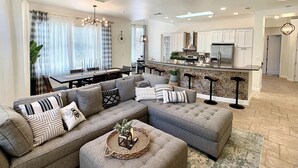 A modern living room with a sectional sofa, a dining area, and a kitchen with white cabinets and a tiled backsplash. - New Vista | Chef Kitchen, Golf Course Views (San Antonio)