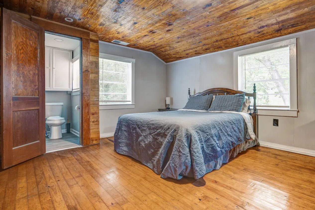 Un dormitorio con techo de madera, una cama con manta azul, dos ventanas y una puerta que da al baño.