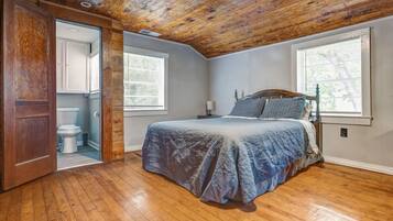 Un dormitorio con techo de madera, una cama con manta azul, dos ventanas y una puerta que da al baño.
