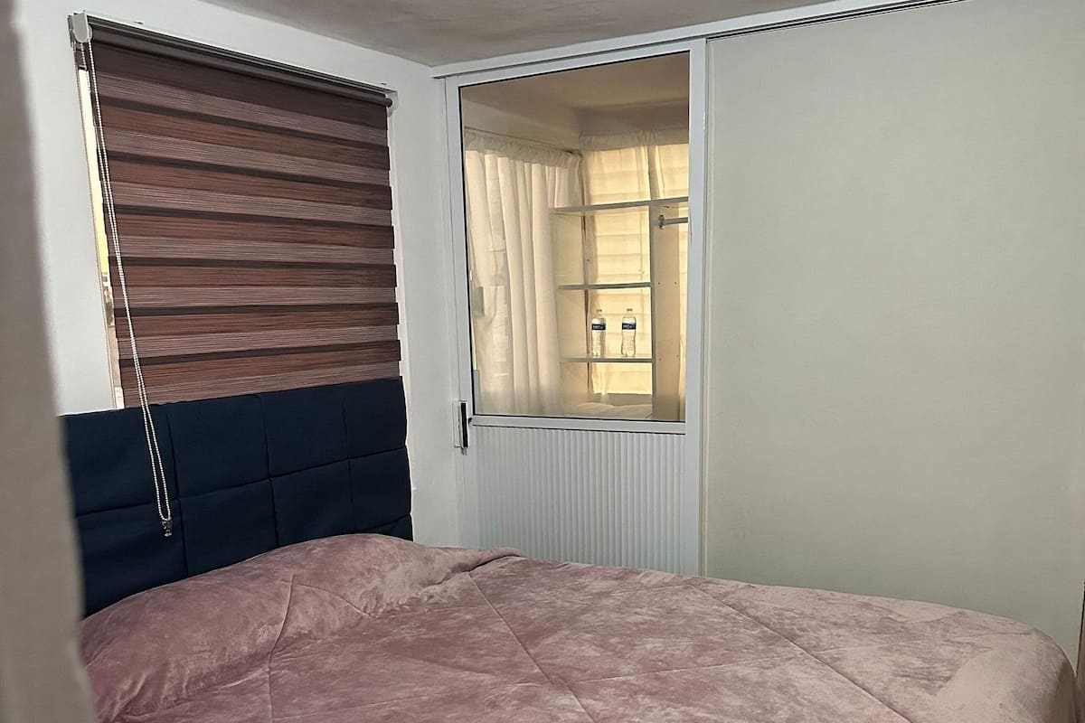 Un dormitorio con cama, ventana con cortinas, radiador y luz de techo.