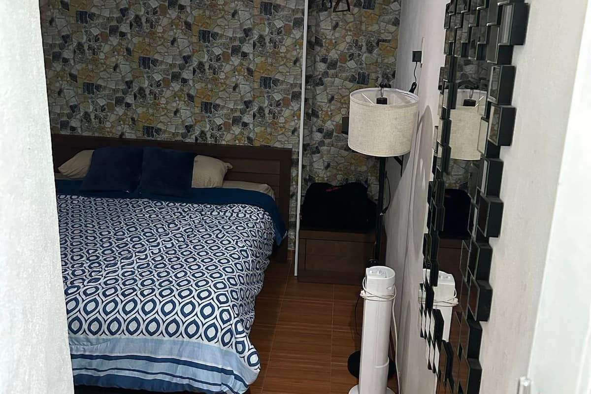 Una habitación pequeña y estrecha con una cama, una lámpara fijada a la pared y un espejo decorativo.