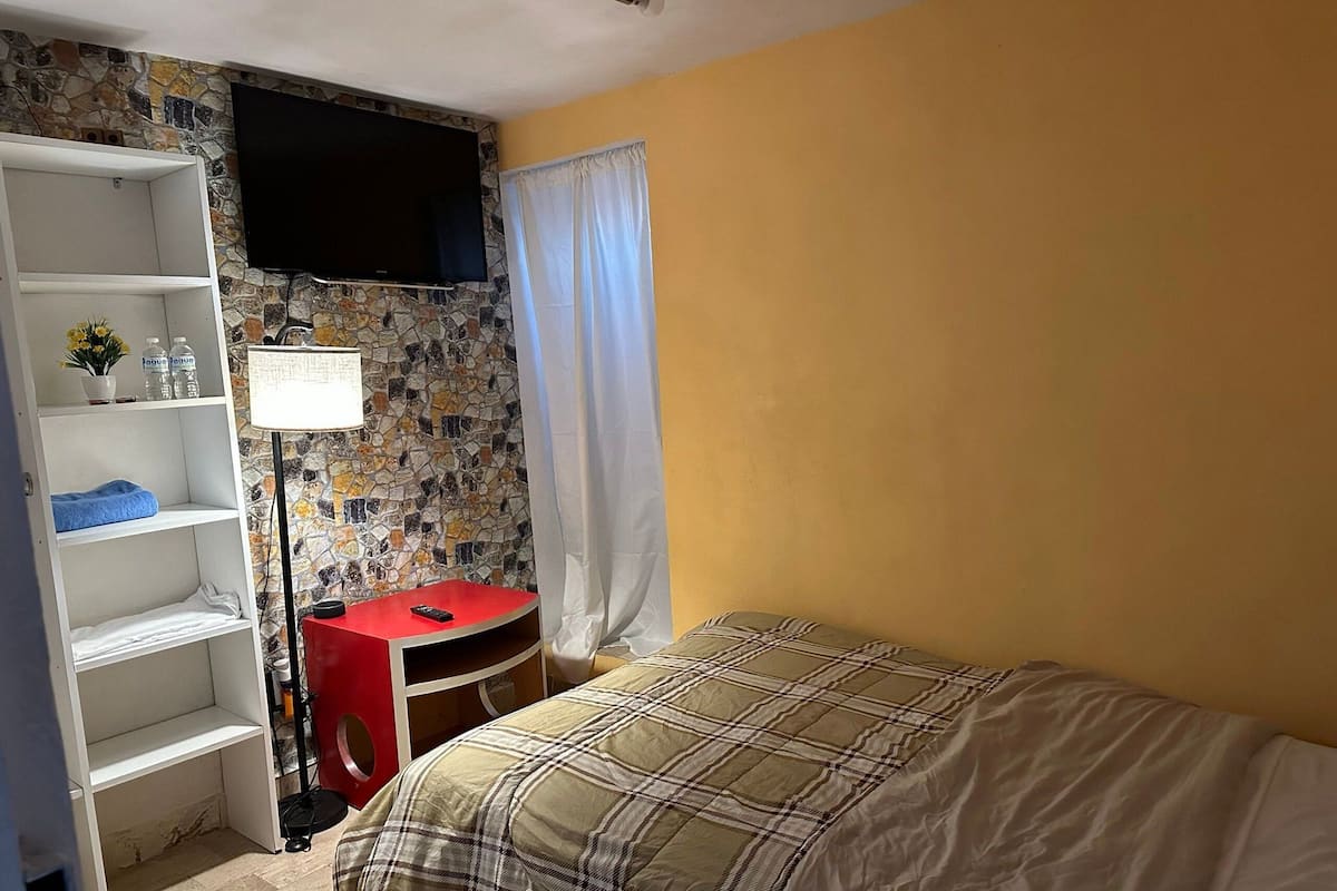 Un dormitorio con una cama, un televisor instalado en una pared de piedra, un escritorio rojo y un estante blanco.