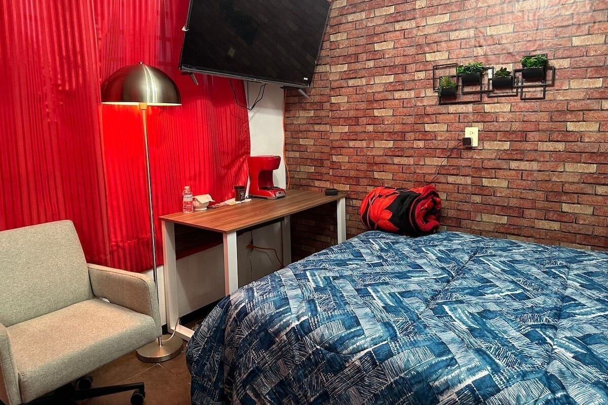 Un dormitorio con cama, escritorio, silla, televisor y pared de ladrillo adornada con plantas en macetas.