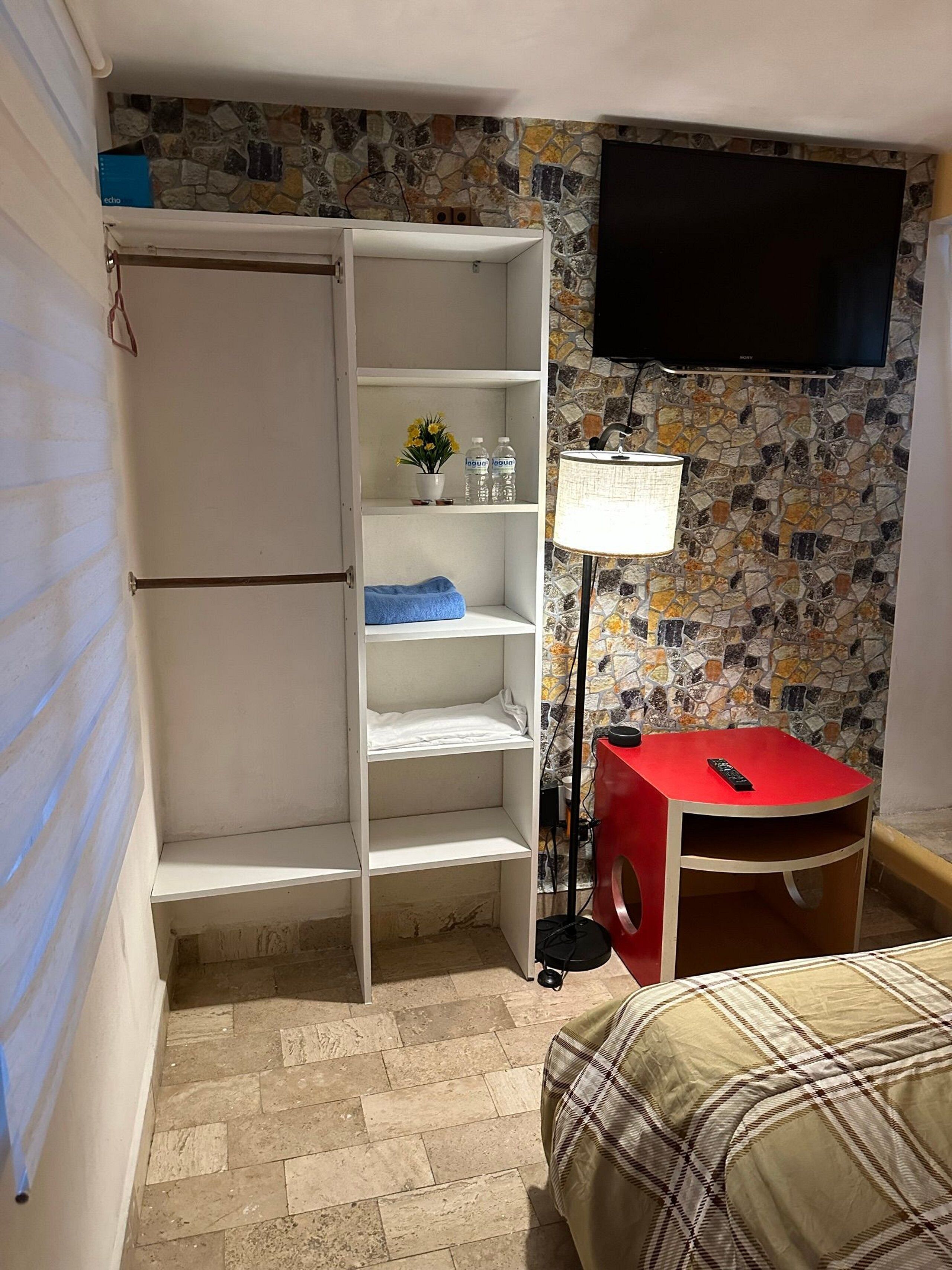 Ein kleiner, kompakter Raum mit einem weißen Kleiderschrank, einem roten Hocker, einem Bett, einem an der Wand befestigten Fernseher und einem gefliesten Boden.