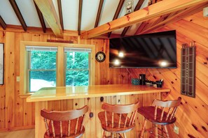A wooden interior with a bar area, two chairs, a television, and a clock. - Cozy Chemo Pond Cabin w/ Dock & Waterfront Views (Eddington)