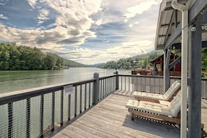 A wooden deck with lounge chairs overlooking a river, surrounded by greenery and a cloudy sky.