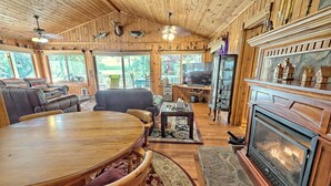 A cozy cabin interior with a wooden dining table, leather sofas, a fireplace, and large windows offering a view of the outdoors. - T Landing Top & Bottom Units (Clayton)