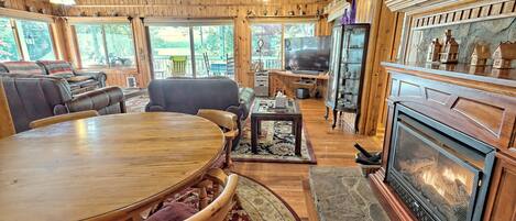 Interior acogedor de una cabaña con una mesa de comedor de madera, sofás de cuero, una chimenea y ventanas amplias que ofrecen vistas del exterior.