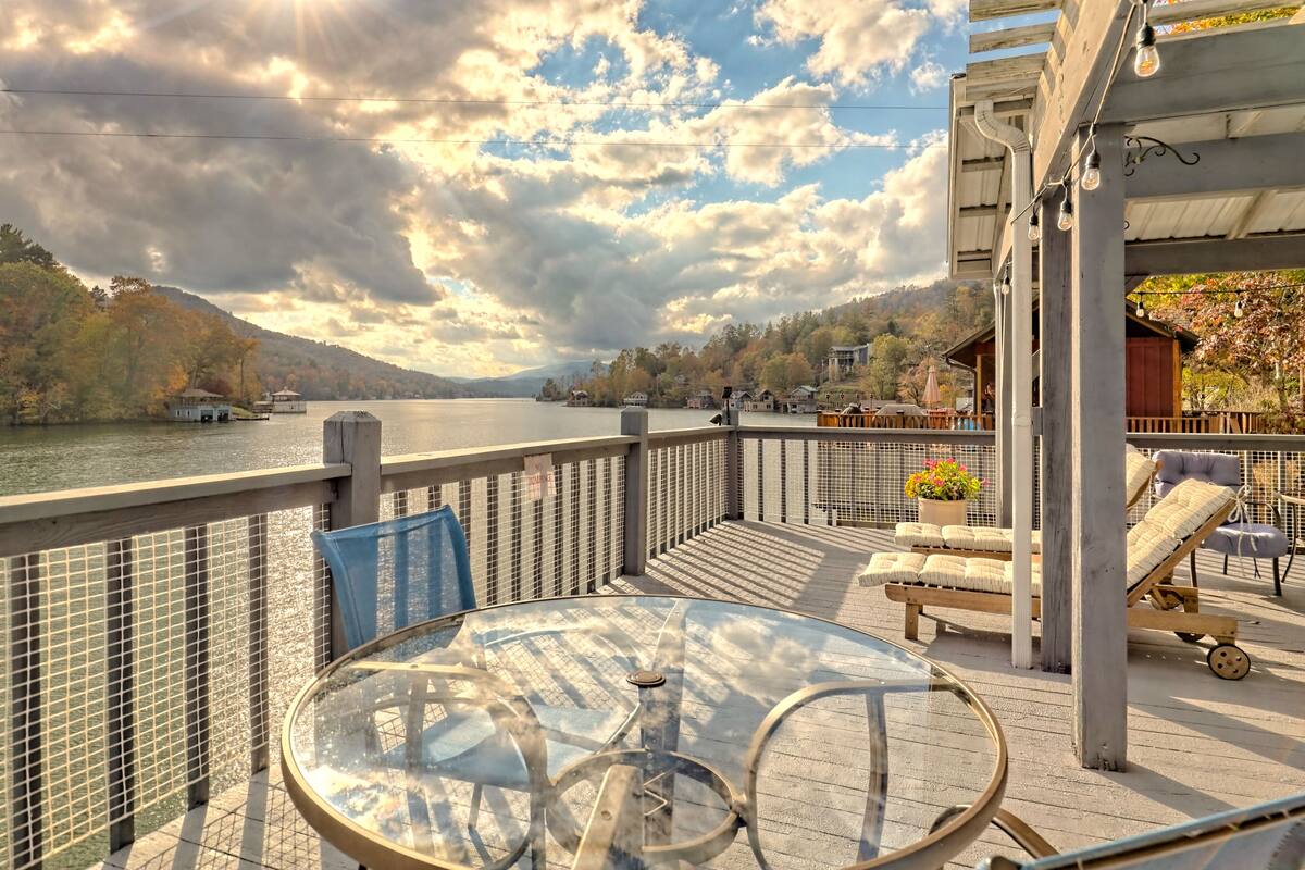 A deck with a glass table and chairs, overlooking a lake with houses and trees in the background.