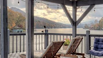 Une terrasse couverte avec deux chaises longues en bois et une chaise bleue, donnant sur un lac avec des montagnes en arrière-plan.